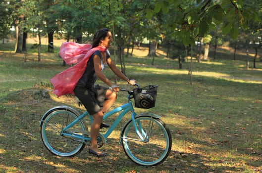 Image of a happy young woman with a pink scarf riding a bicycle in an early autumn park.