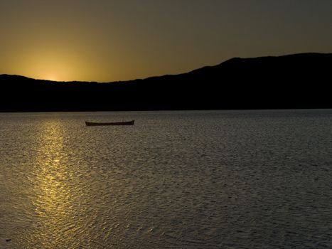 The sun behind the mountains is reflected over the lake surface, beside an abandoned boat.