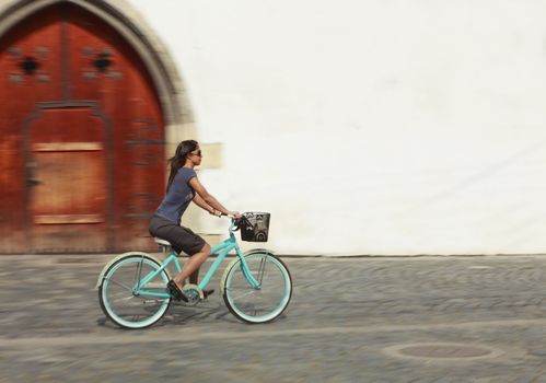 Panning image of a young woman riding her bicycle in a traditional city square in front of an old building with white wall.