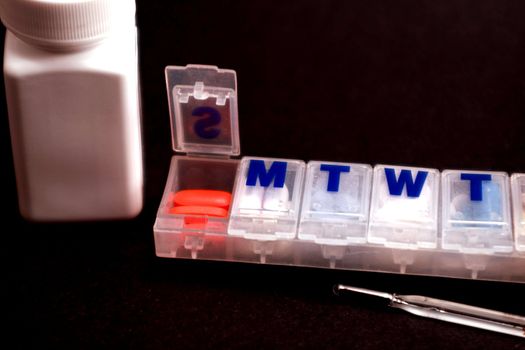 Open pill box, thermometer and medicine bottle on black background.