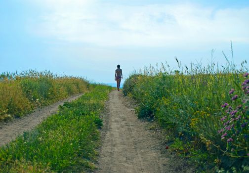 Silhouette of girl in bathing suit, which walks on dirt road overgrown with grass