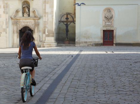Brunette woman riding a bicycle in an old city square in front of a church.