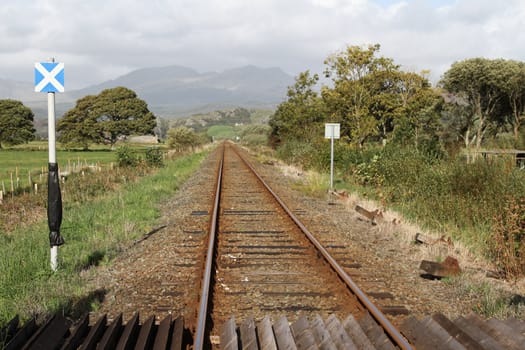 Single track rail-line with crossing and sign in a rural setting with trees, fields and mountains.