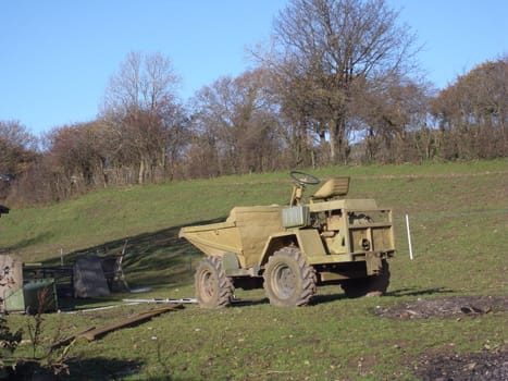 Old green dumper truck in a field with a hedge, with some scrap wood scattered around.