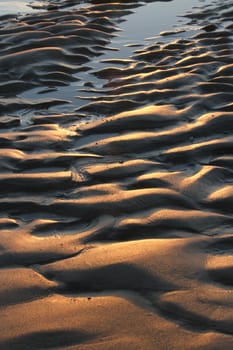 Sand textured with abstract patterns of waves, lines and ruts highlighted by red light and shadows