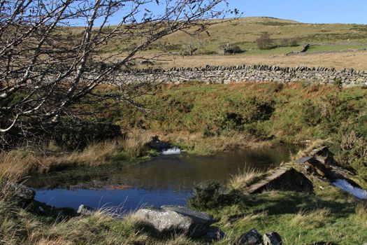 Farm water storage dam with weir amoungst moorland with stone wall and trees.