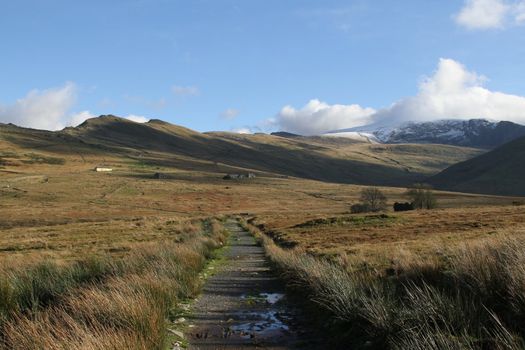 Footpath leading into national park land with distant snowy mountains under cloud, abandoned buildings are dotted across the land.