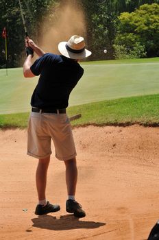 young sporting adult man playing golf shot out of the sand bunker hazard onto the green with the ball visible