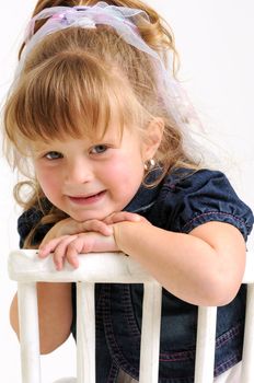 young pretty blond girl with blue dress sitting on a white chair and smiling