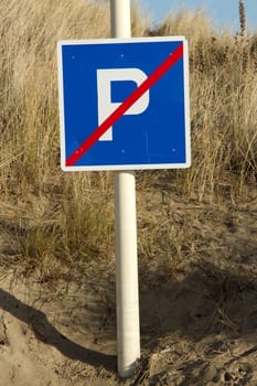 No parking sign, square shaped with the letter P in white on a blue background with a diagonal red line. The sign is attached to a poll and situated in sand with grasses.