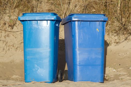 Two blue wheelie bins standing side by side on sand with grasses in the background.