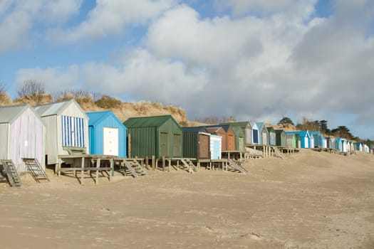 A row of beachhuts, multicoloured, with steps and boardwalks on a beach backed by a sand dune, blue sky and clouds.