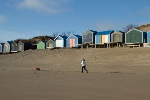 Walker with dog on a beach with a row of beach huts backed by a sand dune and blue sky.