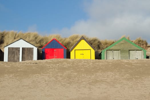 Multi-coloured beach huts with sand banked up against the doors, the scene is backed by a dune with blue sky and cloud.