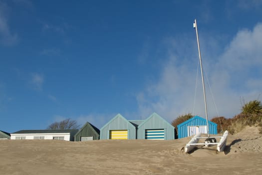 Small yacht, catamaran, in front of beach huts banked up with sand, a blue sky with cloud above.