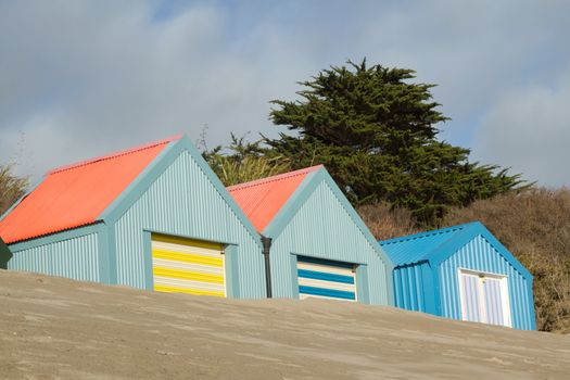 Corrugated metal beachhuts, multicoloured with banked sand and a tree in the background.