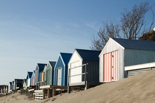 Beach huts, multicoloured on a sand dune,  extend into the distance with a blue sky.