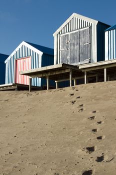 Footprints lead up a sand dune to beach huts on raised platforms, a blue sky backs the scene.