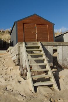 Beach-hut made of metal with wooden doors on a raised wooden platform with a ladder from the beach, a blue sky makes the back-drop.