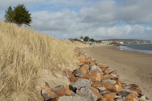 Rocks used for sea defences protecting the sand dunes on a beach with a sea-side village in the distance.