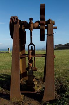 Vintage water pump, rusting metal stand and mechanical components, set in a green grass field with a blue sky.