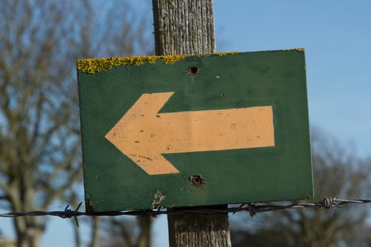 Sign showing an arrow pointing to the left on a green board with lichen attached to a post with barbed wire against a blue sky.