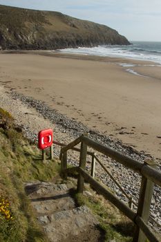 Stone step public footpath with hand rail containing life saving equipment box in red leads to a beach with sand sea and cliff.