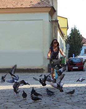 Young woman riding a bicycle in an old city square full of pigeons. 