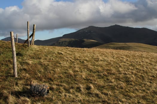 Mountain landscape with grass and undulating ground, fence posts with rusted wire line the side and a coil of new barbed wire lays on the floor.