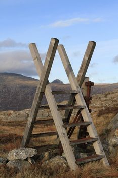 A wooden style, with steps over a wire fence with a post in a mountain location and blue sky.