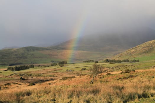 A rainbow forms under a rain cloud in rural fields and trees surrounded by hills.