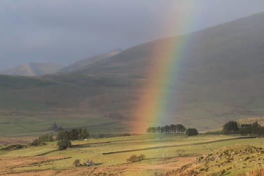 A rainbow forms under a rain cloud in rural fields and trees surrounded by hills.