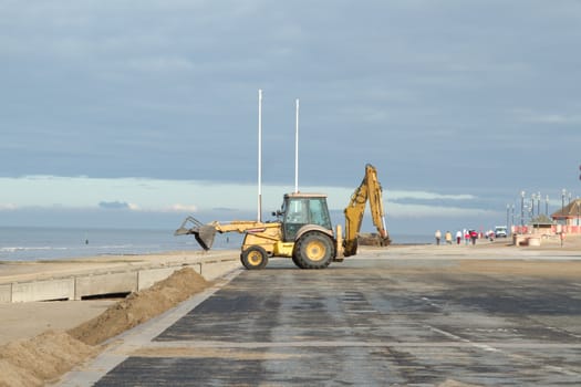 A yellow digger empties sand on the beach from the seafront path, walkers in the distance.