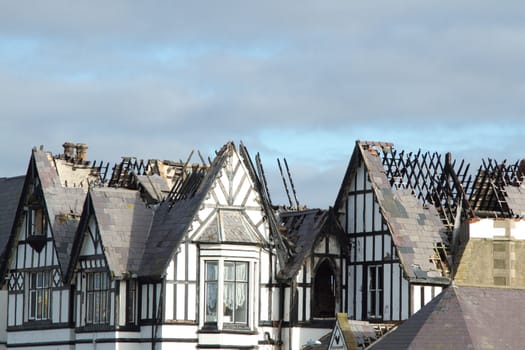 A burnt out building in tudor style with the roof seriously damaged with missing slates and exposed charred rafters.