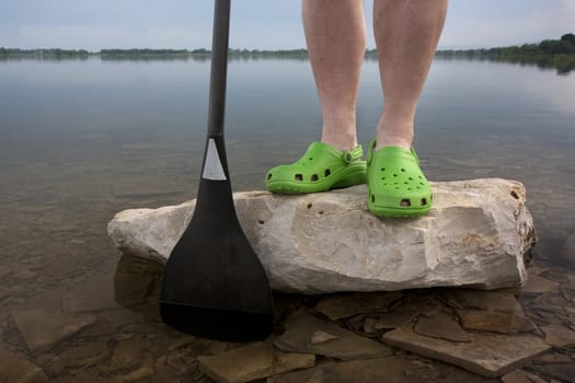 paddler feet in bright green clogs, wide carbon fiber canoe paddle for outrigger racing against lake with a rocky bottom