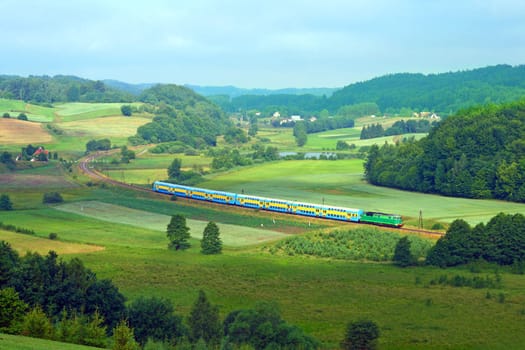 Hilly landscape with a railway line, long train, lake and forests during the summer day