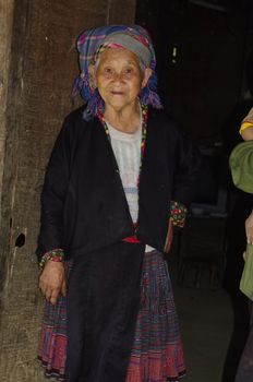 Grandmother Hmong flower on her doorstep. She wears her traditional costume .Colorful dress, apron and black jacket and a multicolored wool scarf on.