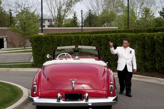 A sharp dressed man whistling and pointing walking next to a classic convertible car.