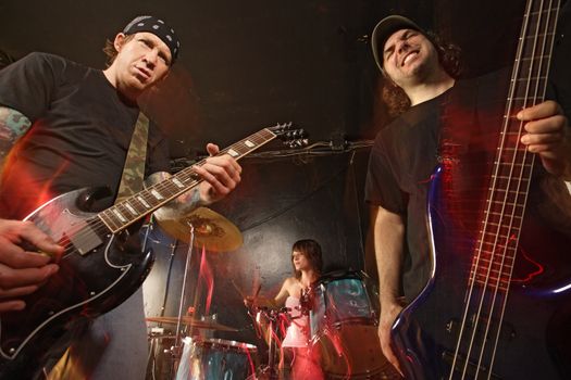 Band playing on a stage. Guitarist, bassist and female drummer. Shot with strobes and slow shutter speed to create lighting atmosphere and blur effects. Slight motion blur on performers.