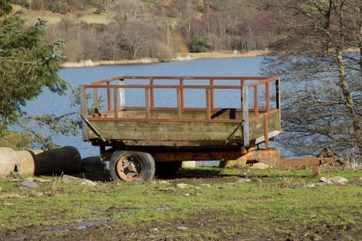 An old trailer with flat tires tow-bar, made from wood and metal on grass and mud with a lake in the background.