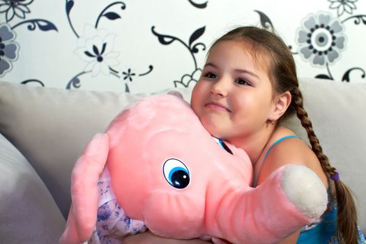 The little girl is sitting on the couch and big hugs plush toy.