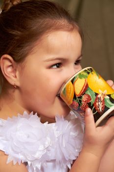 The little girl in a white dress drinks from a large mug.