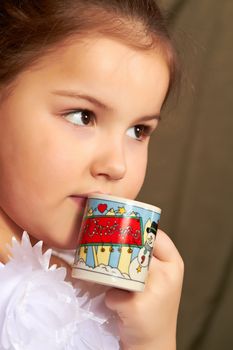 The little girl in a white dress drinks from a small cup.