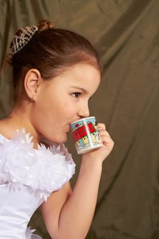 The little girl in a white dress drinks from a small cup.