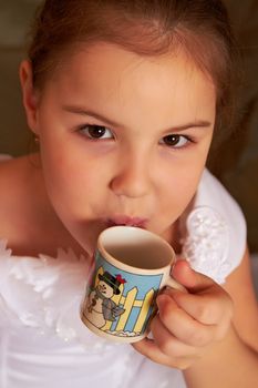 The little girl in a white dress drinks from a small cup.