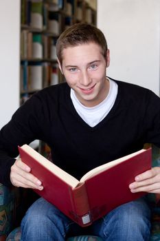 A young college aged man reading a book at the library with a smile.