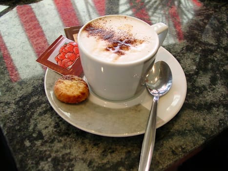 This is a nice hot cup of café mocha coffee with sprinkled chocolate on a saucer with a spoon and cookie placed on a granite table.