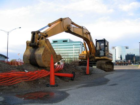 This is an excavator in a work area, waiting to start digging into a new construction zone with buildings in the background.