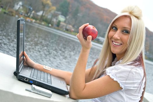 A beautiful young blonde woman in a mobile business setting eating a red apple while using her laptop notebook computer.