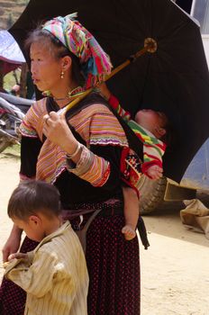 Woman flowered Hmong .Near her ,her son and she carries in her back her baby .She came from far in the mountains this Saturday at Can Cau market shopping